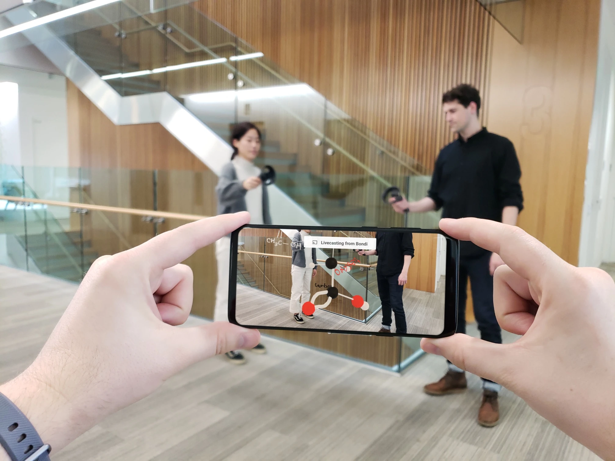 A phone screen reveals virtual molecules in a shared XR scene with two people holding controllers